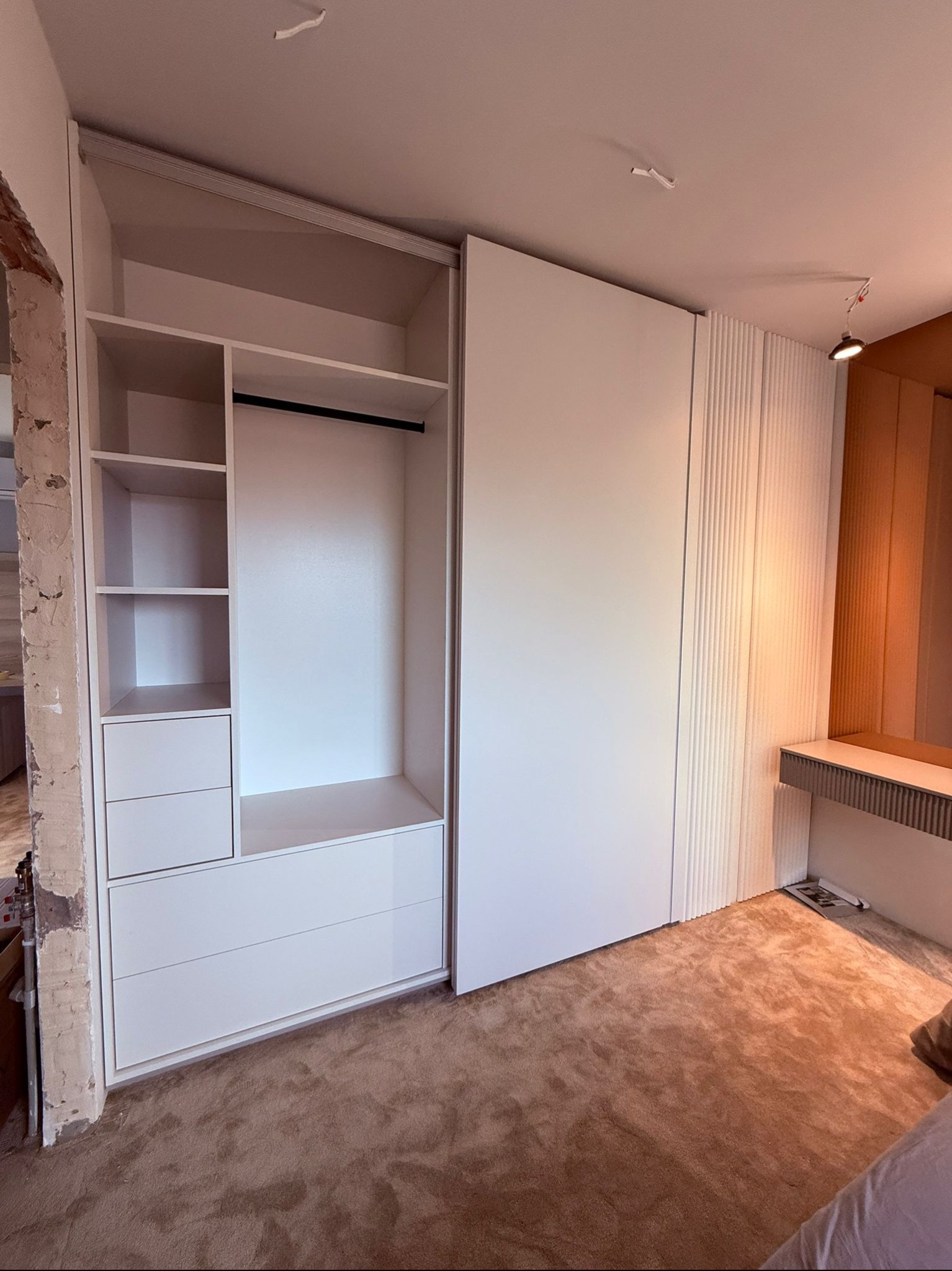 Biała szafa wnękowa z przesuwnymi drzwiami, widoczne półki i szuflady, surowy mur przy szafie, dywan w odcieniach brązu, oświetlenie punktowe.