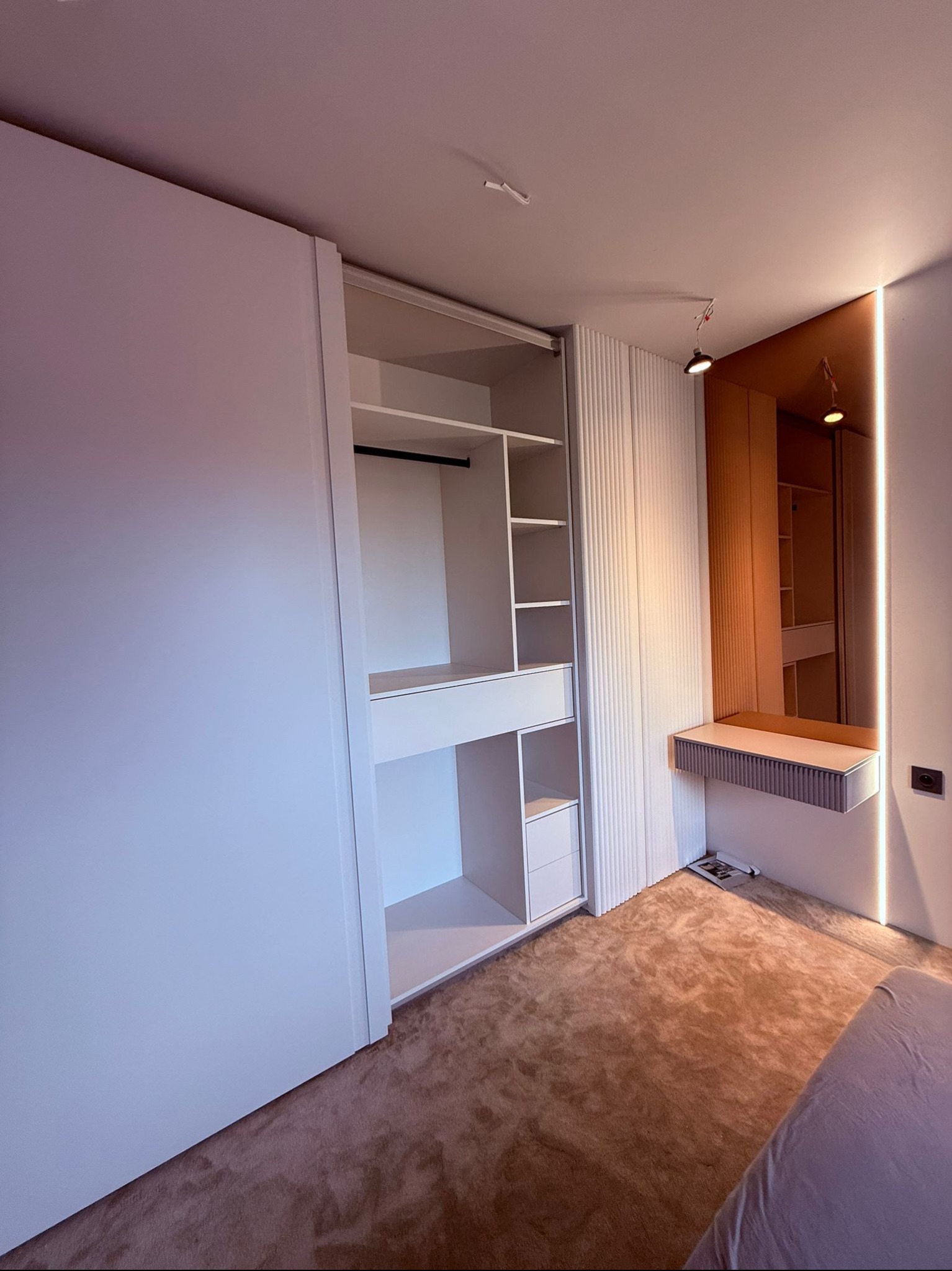 Nowoczesna szafa wnękowa z przesuwnymi drzwiami, oświetleniem LED i toaletką wkomponowana w aranżację sypialni z beżową wykładziną. Minimalistyczny design.