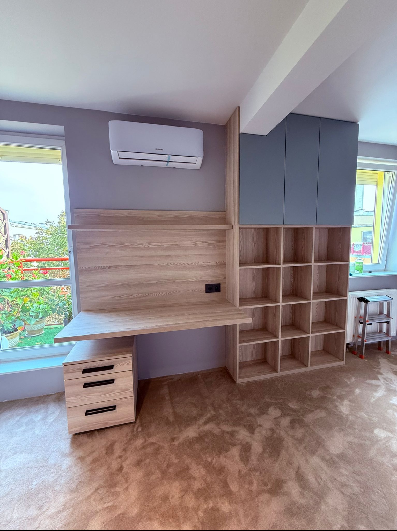 Jasne drewno biurka z szafkami i półkami, szare szafki wiszące, widok na okno z zielenią, brązowa podłoga. Nowoczesne meble do pracy w domu.