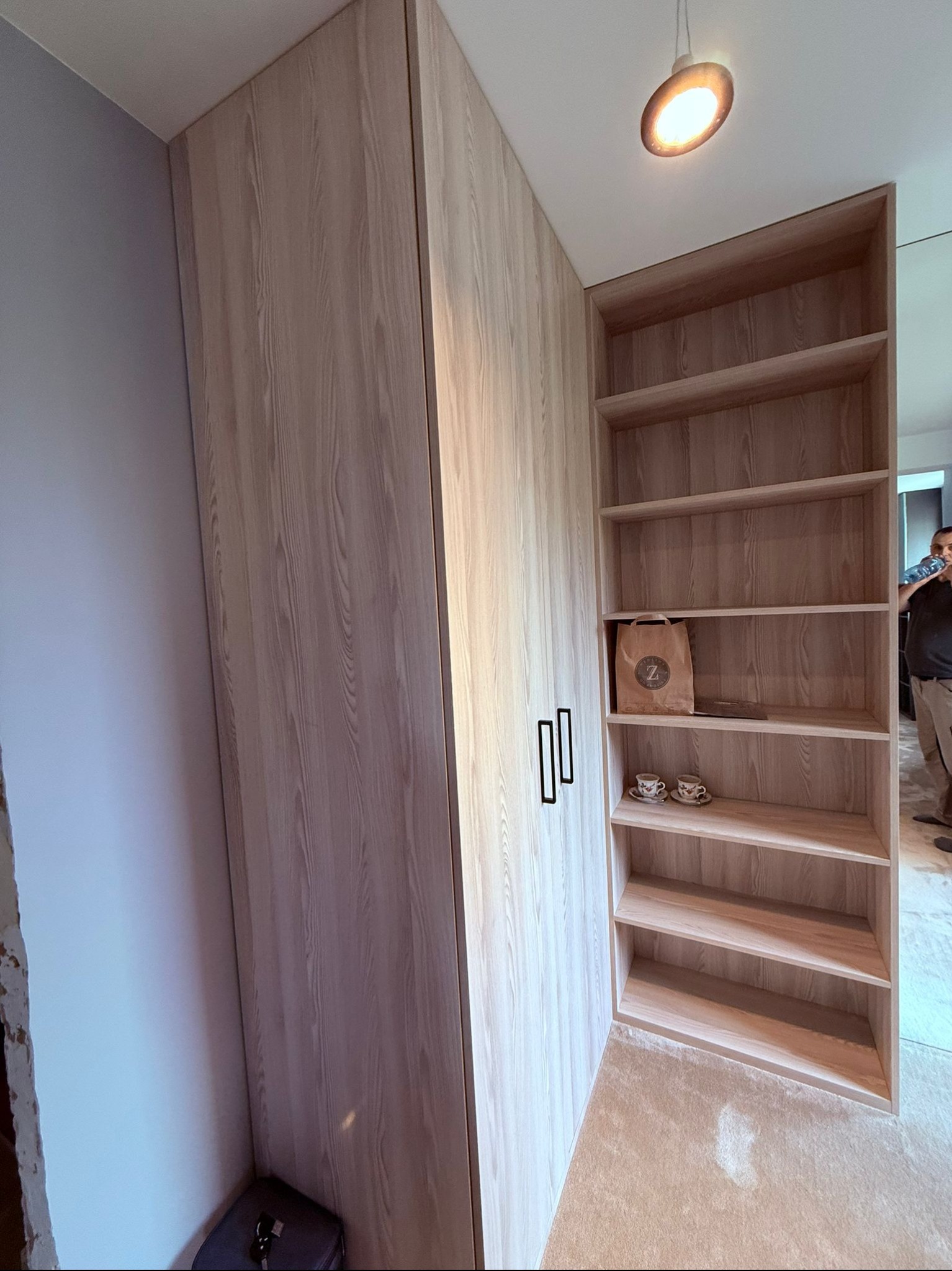 Jasna, drewniana szafa wnękowa z otwartymi półkami, uzupełniona o torbę i filiżanki. Minimalistyczne, czarne uchwyty. Oświetlenie sufitowe.