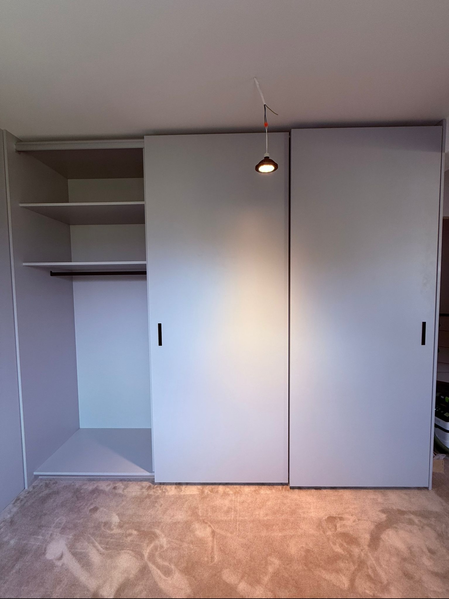 Jasnoszara szafa przesuwna z czarnymi uchwytami, oświetlona punktowo, na tle beżowej wykładziny. Widoczny otwarty moduł z półkami i drążkiem.