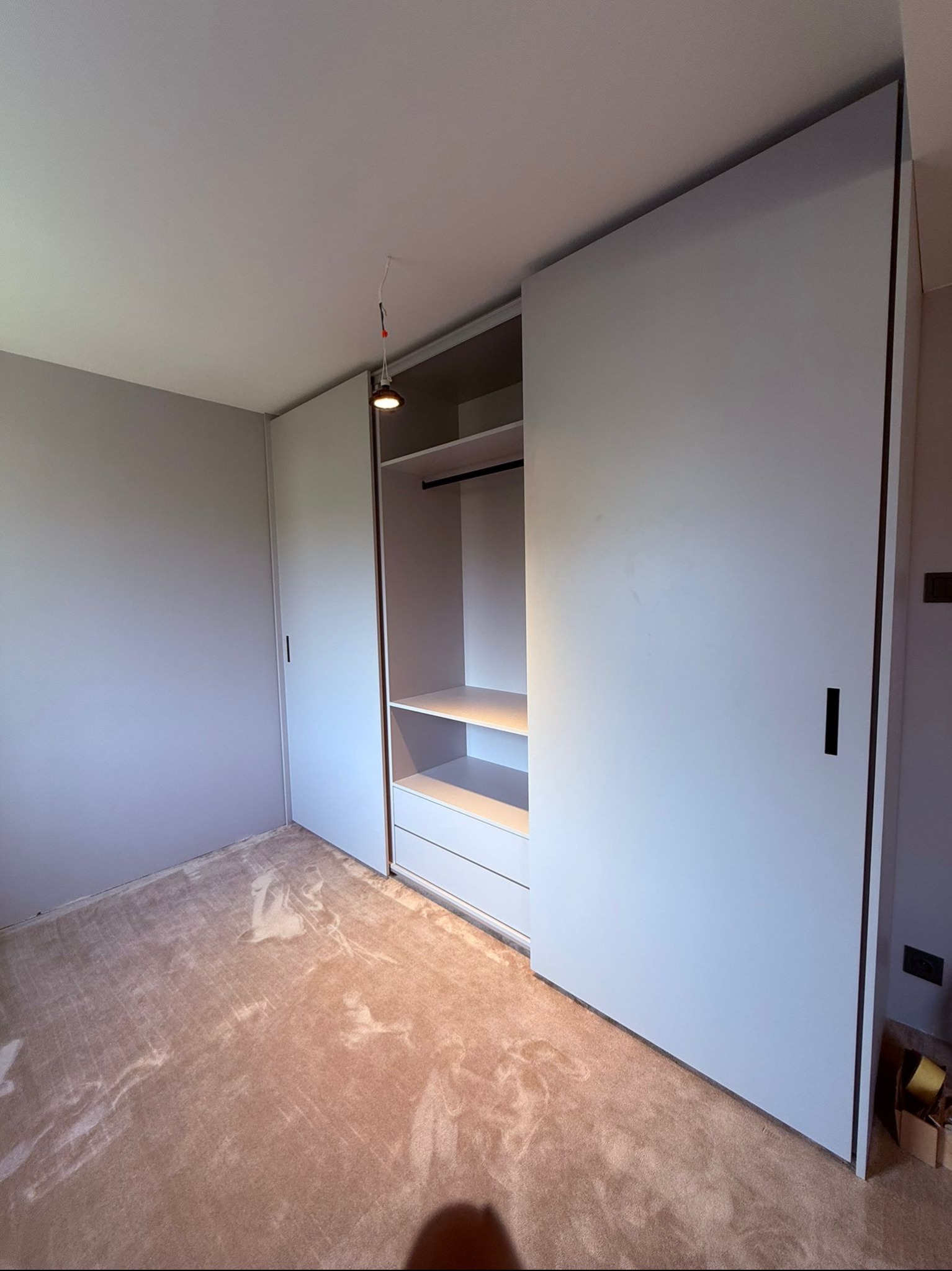 Jasnoszara szafa wnękowa z przesuwnymi drzwiami i otwartą częścią z półkami i drążkiem, widoczna w pokoju z beżową wykładziną. Minimalistyczny design.