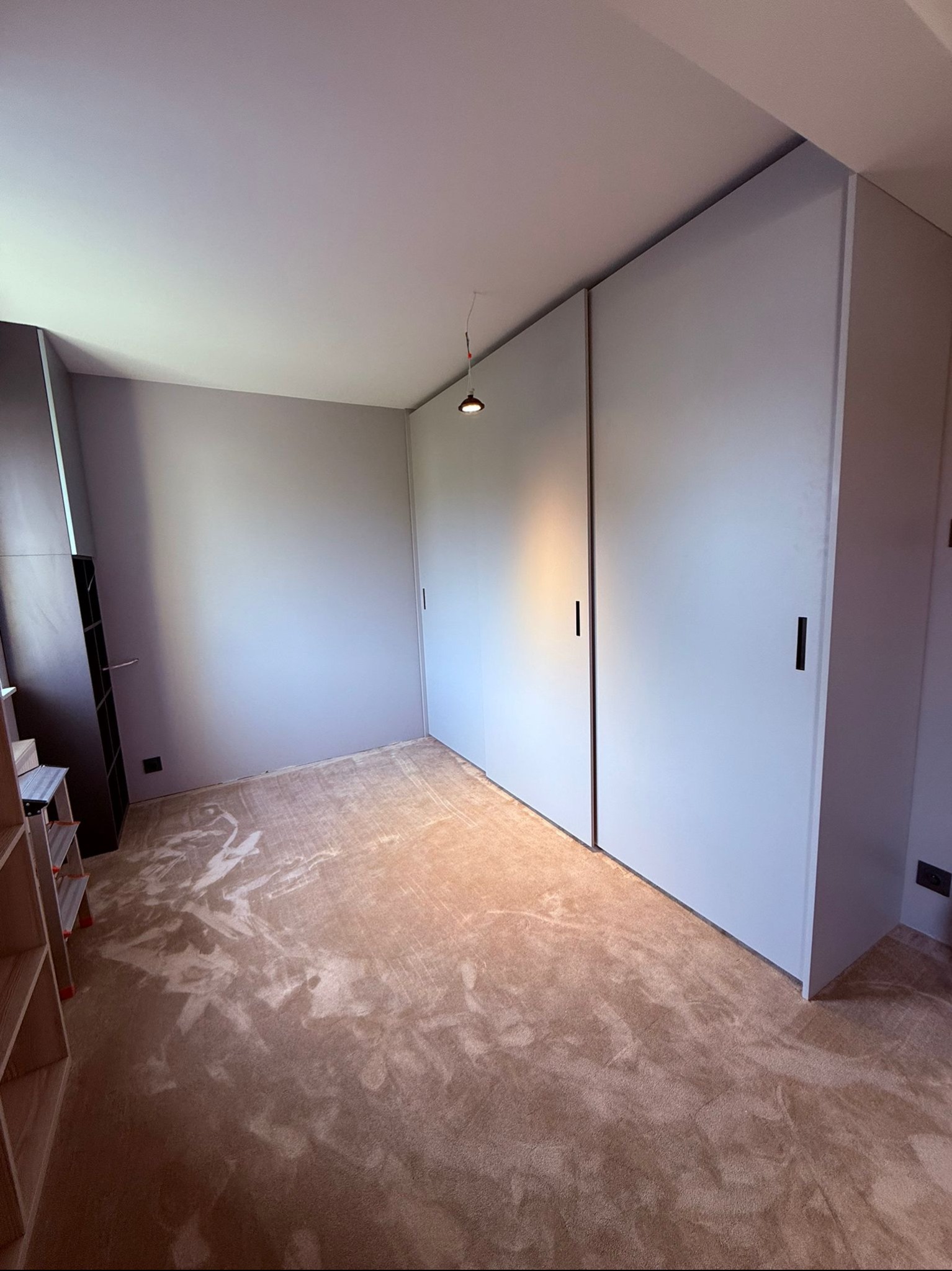 Jasne, minimalistyczne wnętrze z dużą szafą przesuwną o gładkich, białych frontach. Na podłodze beżowa wykładzina. W tle regał z ciemnego drewna.