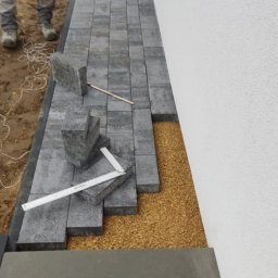 YOKO-BUD - Układanie szarej kostki brukowej przy białej ścianie budynku, widoczne narzędzia pomiarowe i warstwa podsypki.