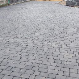 YOKO-BUD - Ułożona szara kostka brukowa na posesji, w tle minikoparka i ubijarka wibracyjna, widoczny fragment ogrodzenia i budynków.