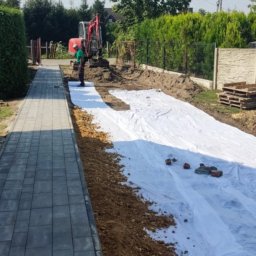 YOKO-BUD - Przygotowanie terenu pod budowę, widoczna wykopana ziemia, biała geowłóknina, pracownik w zielonej koszulce i koparka w tle, obok ułożona kostka brukowa.