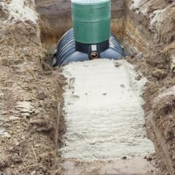 YOKO-BUD - Czarna plastikowa studzienka kanalizacyjna z zieloną pokrywą umieszczona w wykopie, częściowo zasypana piaskiem, widoczny pracownik w tle.