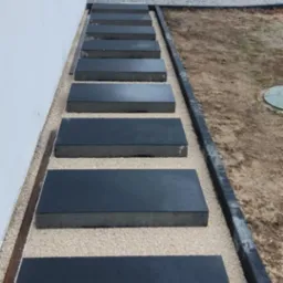 Nowoczesne, prostokątne płyty chodnikowe w kolorze grafitowym, ułożone wzdłuż białej ściany budynku na jasnym żwirze, tworzące ścieżkę. Krawężnik oddziela żwir od ziemi. W tle widoczne stosy...