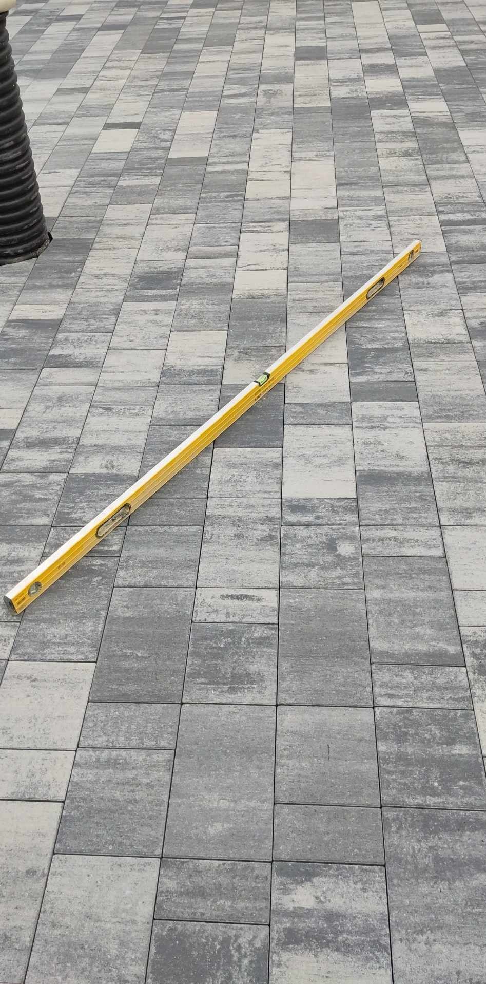 Ułożona szara kostka brukowa z poziomnicą w trakcie sprawdzania poziomu, widoczny element drenażu.