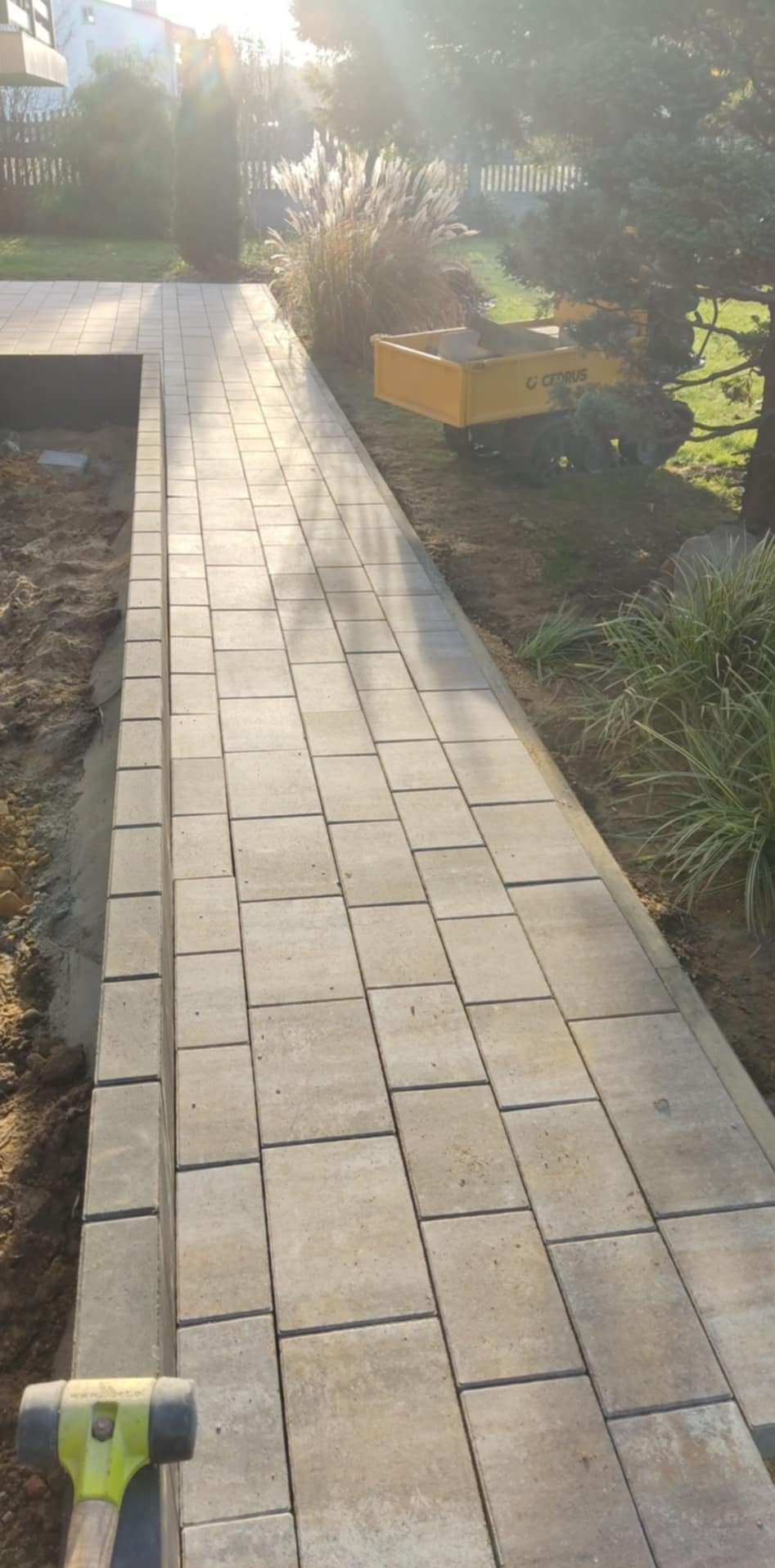 Świeżo ułożona ścieżka z beżowej kostki brukowej w ogrodzie, z widocznym gumowym młotkiem i taczką w tle, pod delikatnym światłem słonecznym.