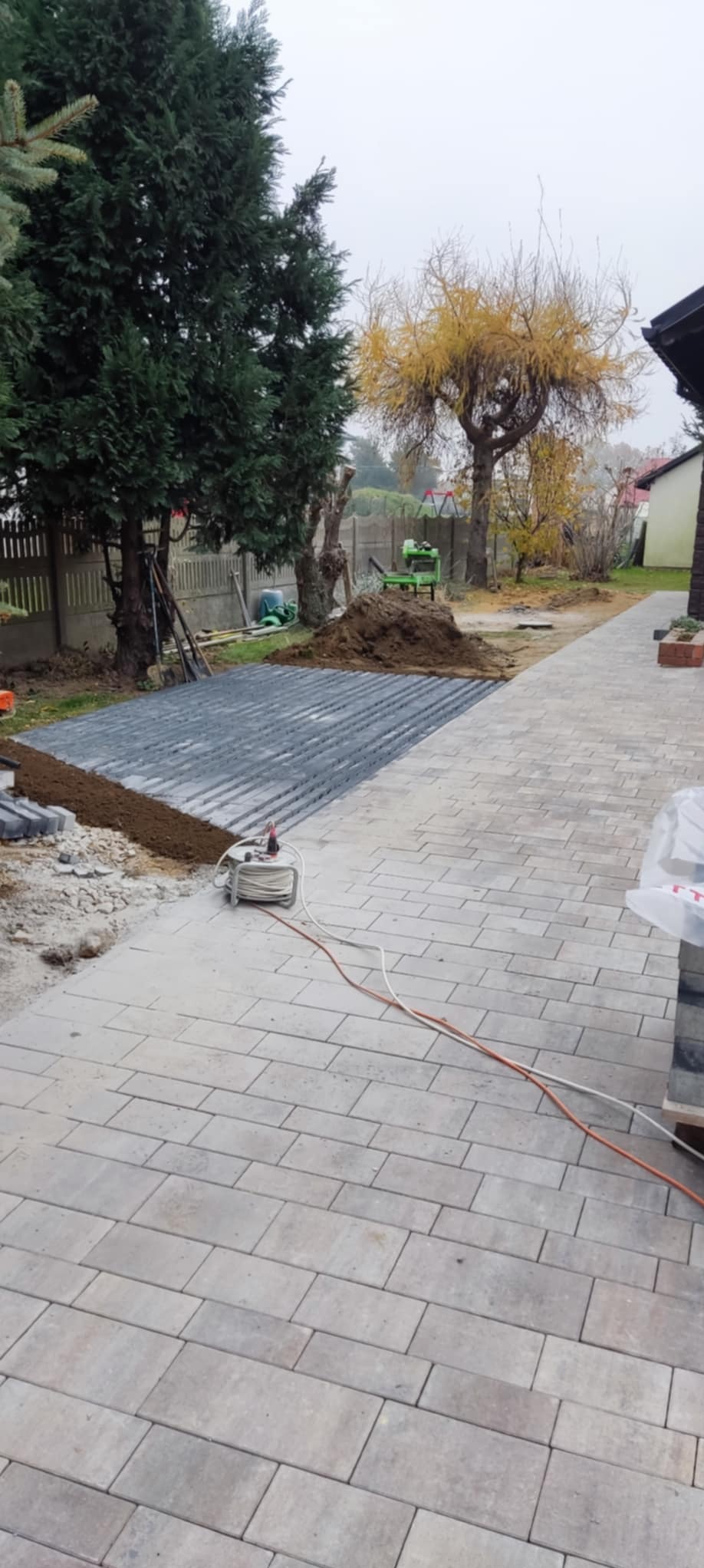 Układanie ciemnoszarej kostki brukowej na przygotowanym podłożu, widoczne narzędzia i kable, w tle drzewa i ogrodzenie.