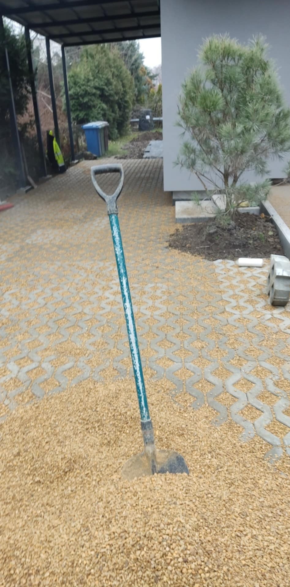 Eko-kratka wypełniona drobnym żwirem, z wbita w podłoże łopatą o zielonym trzonku, widok na podjazd i dom z iglakiem