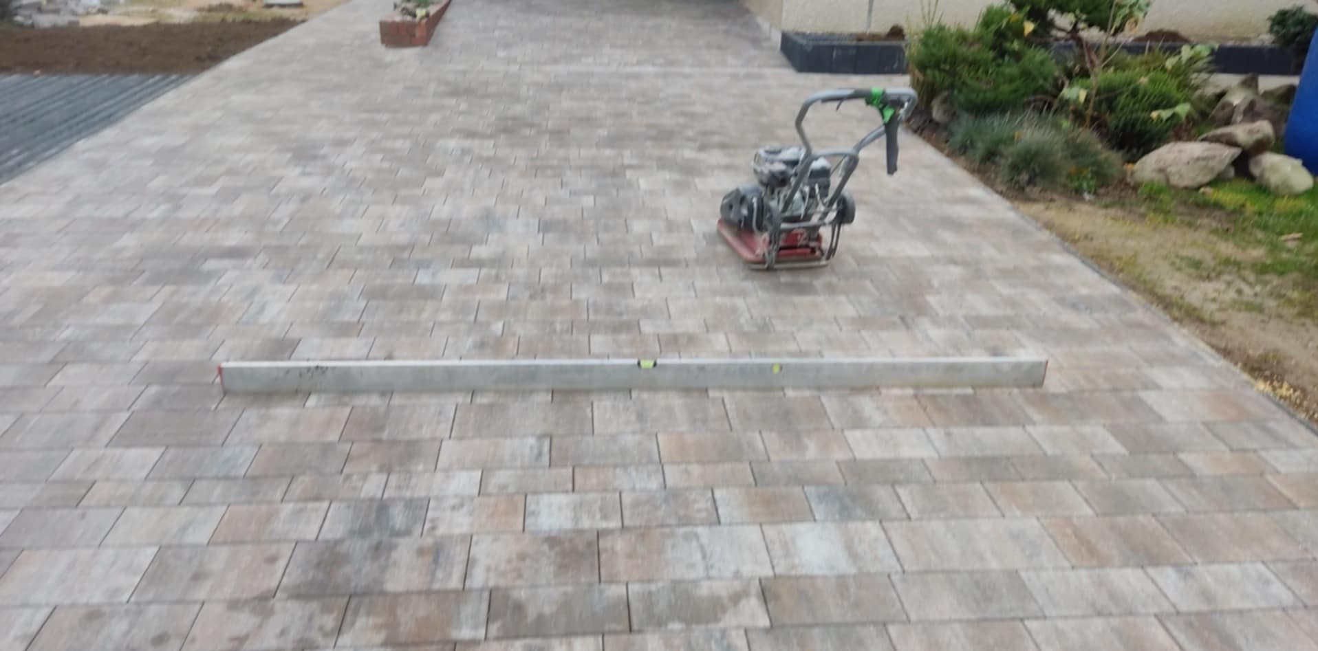 Ułożona, beżowo-szara kostka brukowa z widoczną poziomicą i ubijarką, otoczona zielenią i elementami ogrodu.