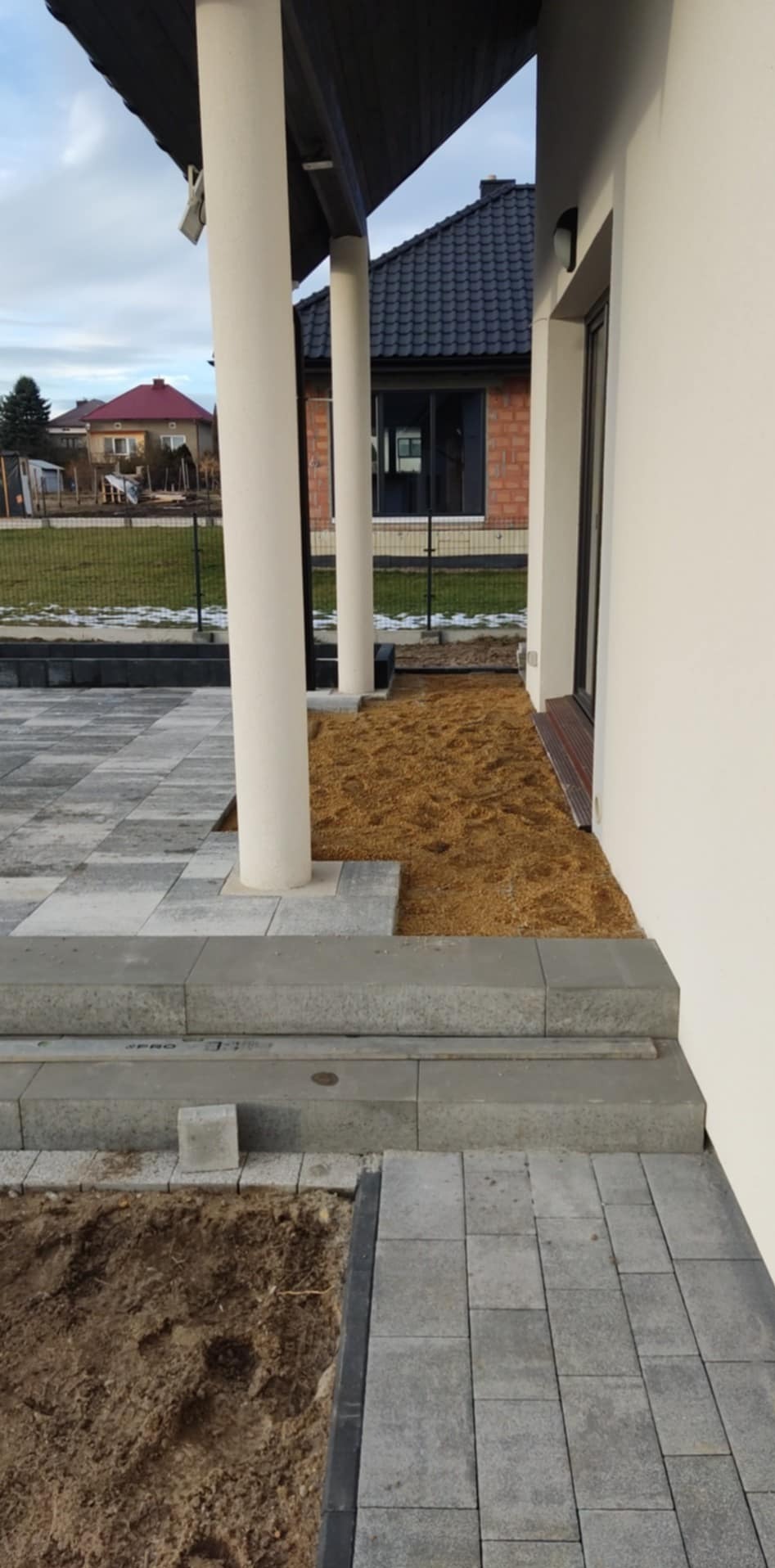 Fragment tarasu z szarej kostki brukowej, widoczne schody wejściowe i fragment rabaty z korą, w tle biała elewacja budynku i kolumny podtrzymujące zadaszenie.