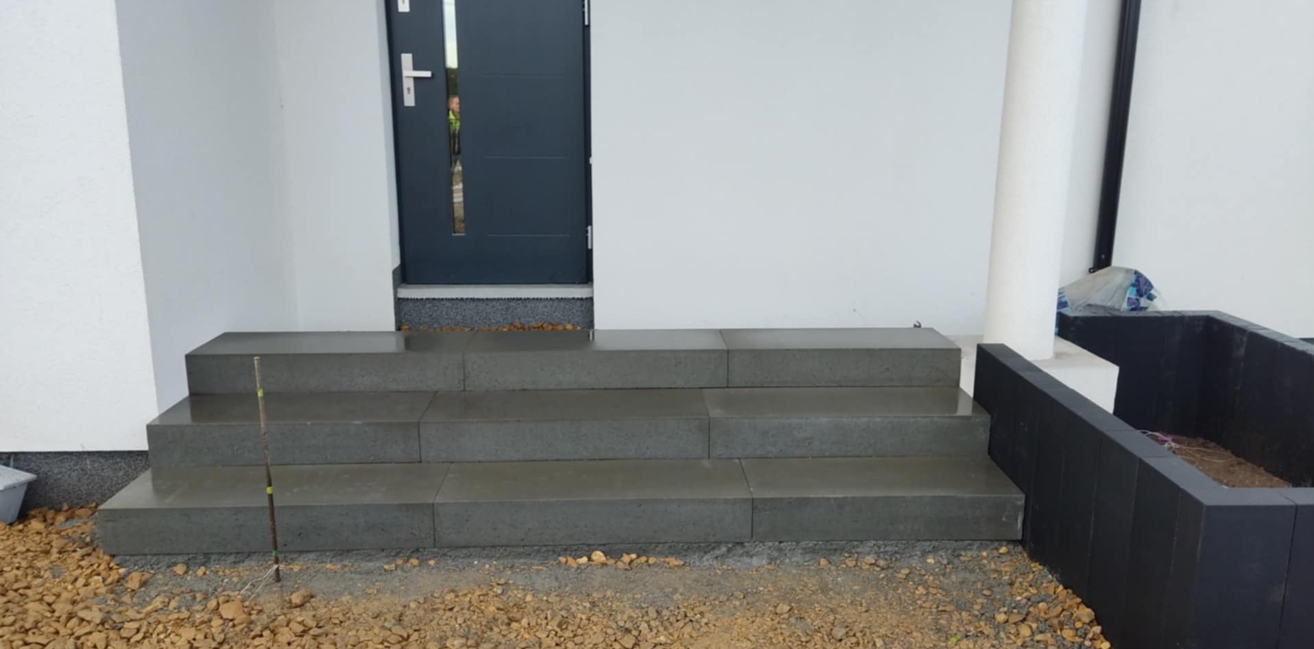 Schody wejściowe wyłożone szarą kostką brukową prowadzące do nowoczesnych drzwi wejściowych, widoczne przygotowane podłoże z kamyków i fragment donicy z ciemnego tworzywa.