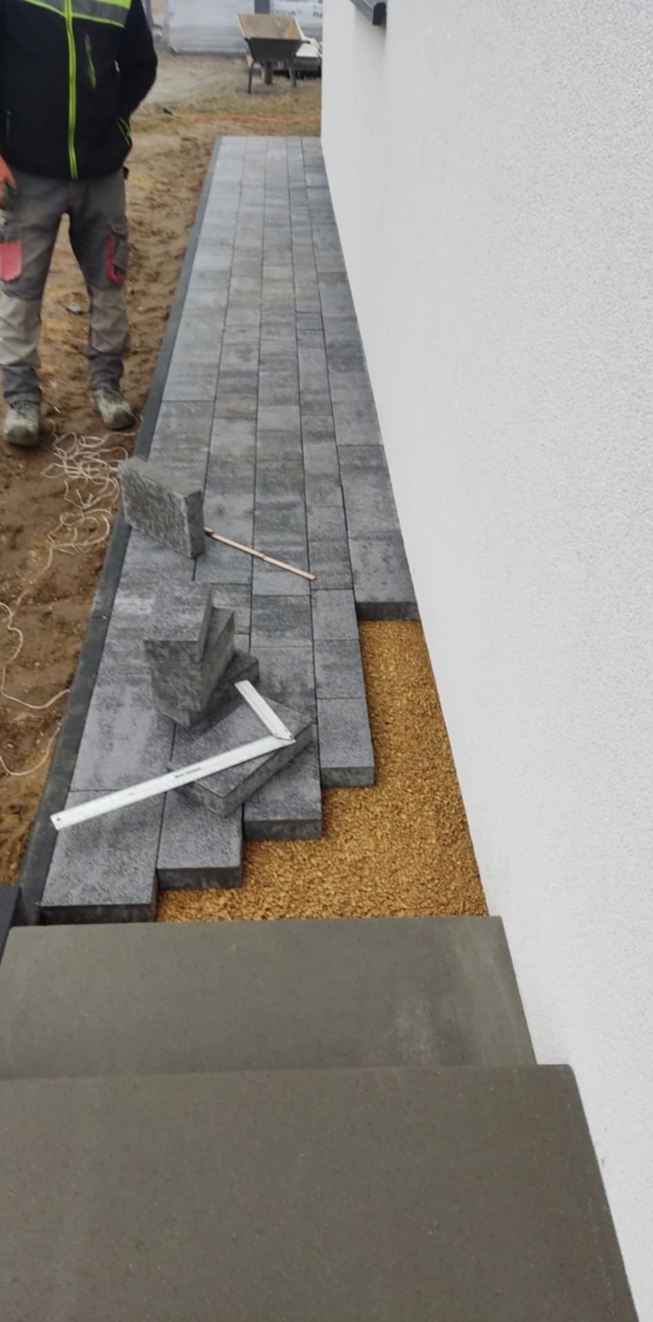 Układanie szarej kostki brukowej przy białej ścianie budynku, widoczne narzędzia pomiarowe i warstwa podsypki.