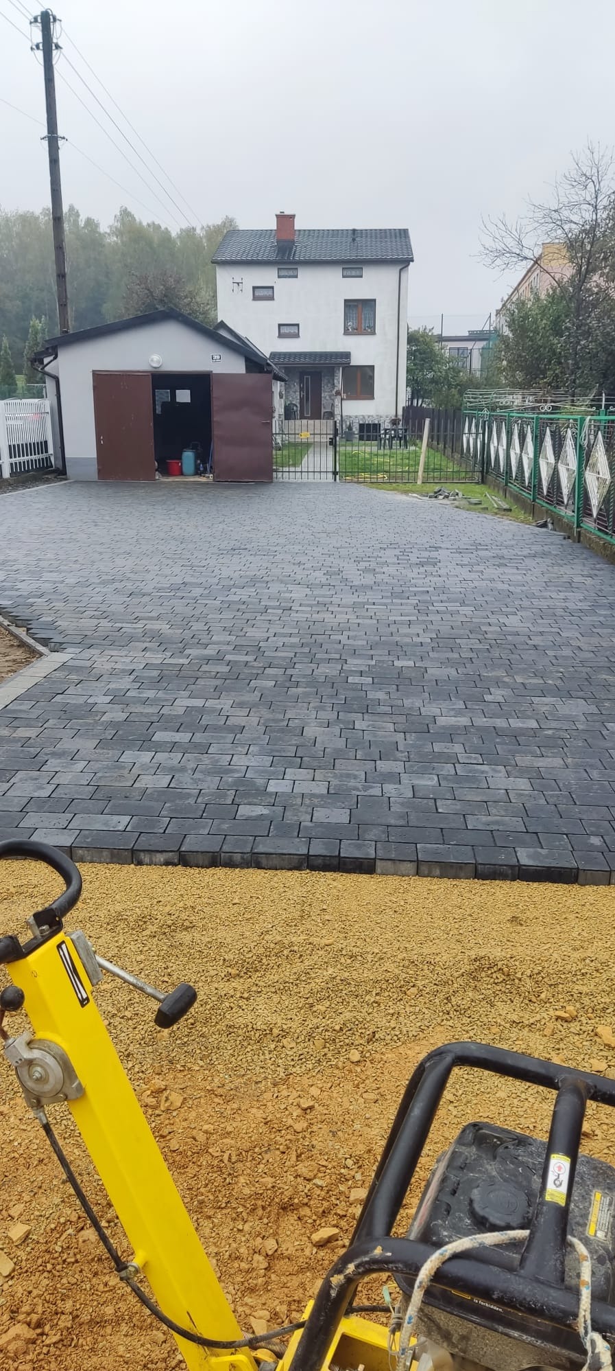 Ułożona, szara kostka brukowa na podjeździe przed domem, widoczna ubijarka w trakcie pracy na podsypce piaskowej.