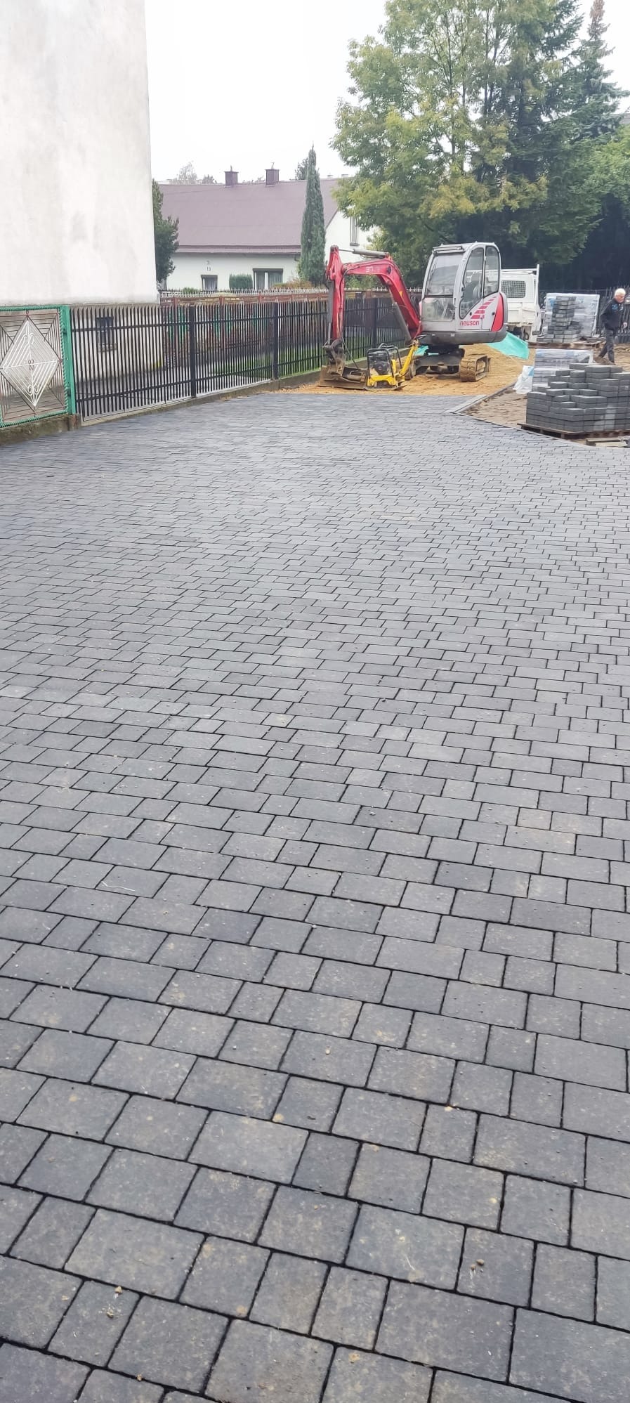 Ułożona szara kostka brukowa na posesji, w tle minikoparka i ubijarka wibracyjna, widoczny fragment ogrodzenia i budynków.