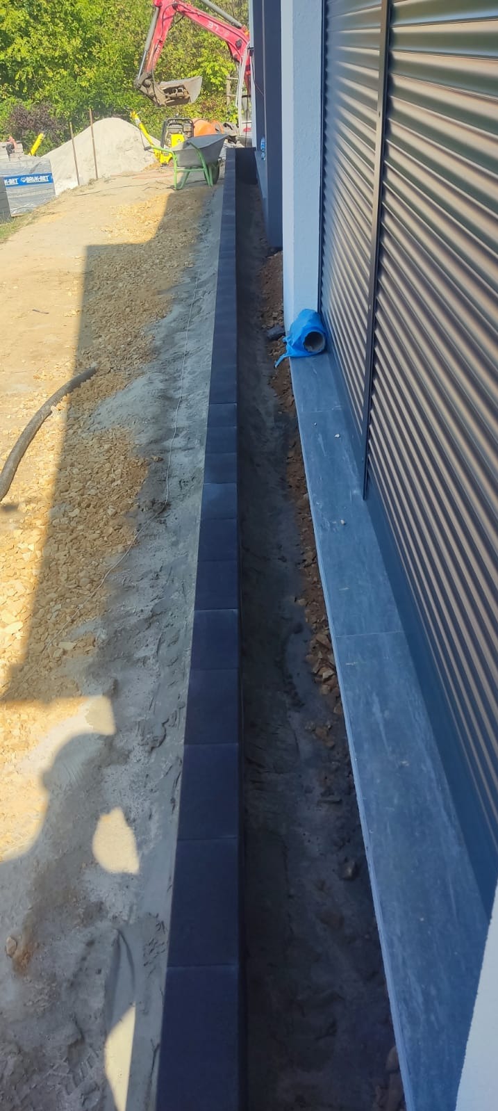 Wykop fundamentowy obłożony rzędem ciemnych betonowych bloczków, widoczny wykopany grunt, rura od pompy, w tle wykop ziemny i mała koparka.