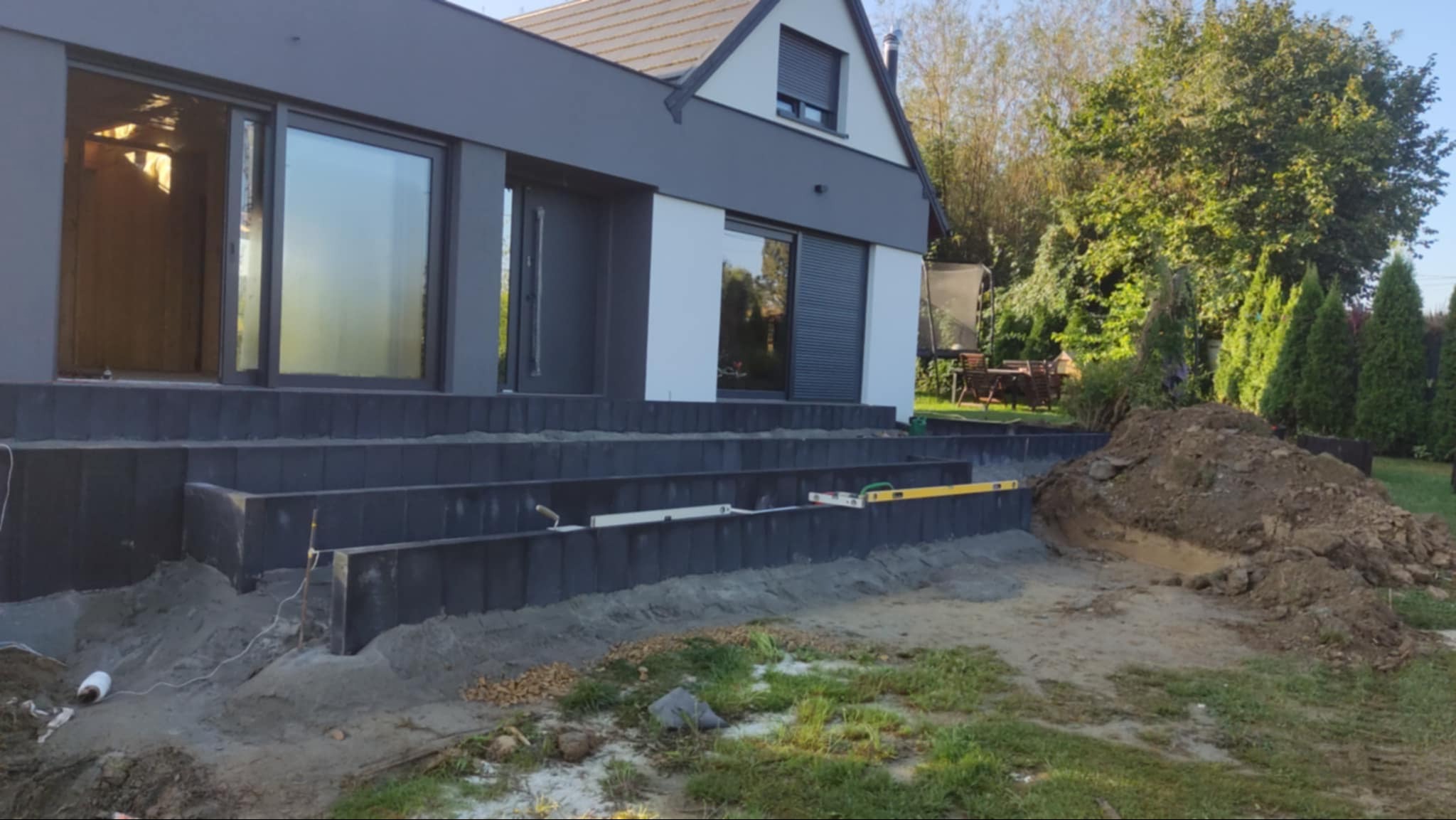 Budowa schodów zewnętrznych z elementów betonowych przy nowoczesnym domu. Widoczne poziomice i nasyp ziemi.