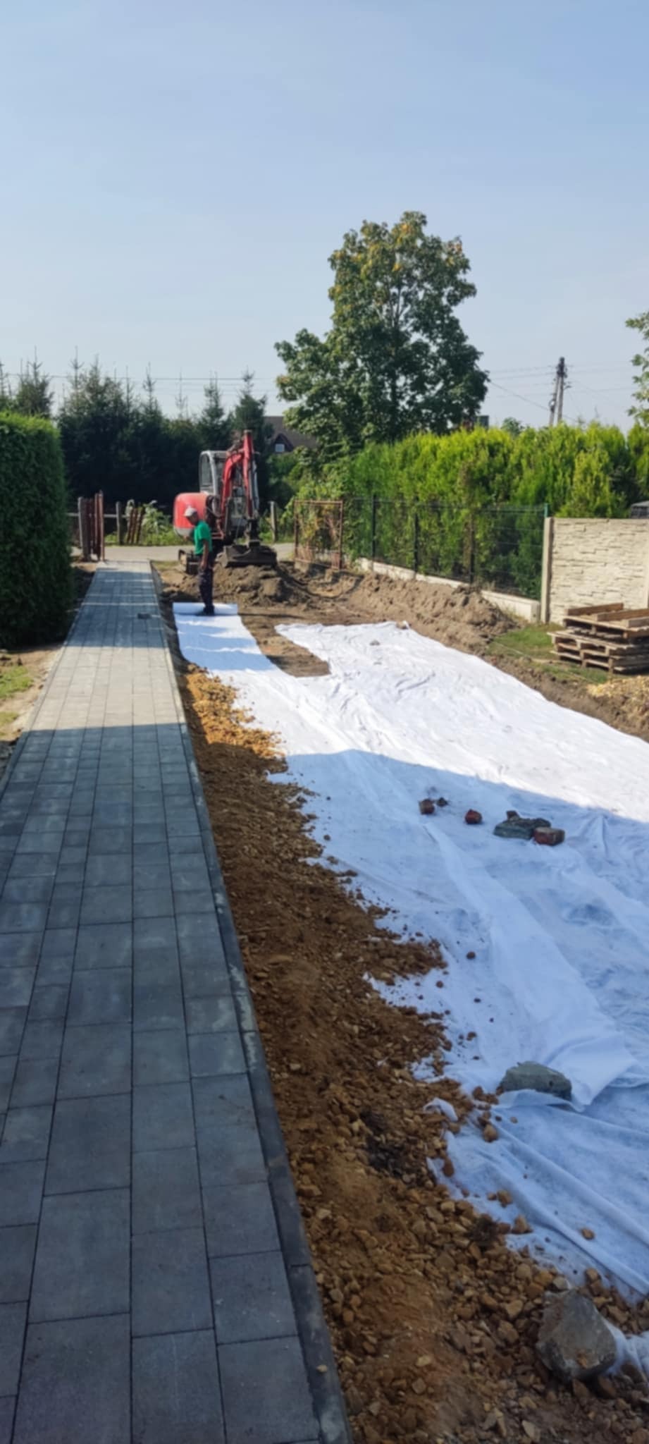 Przygotowanie terenu pod budowę, widoczna wykopana ziemia, biała geowłóknina, pracownik w zielonej koszulce i koparka w tle, obok ułożona kostka brukowa.