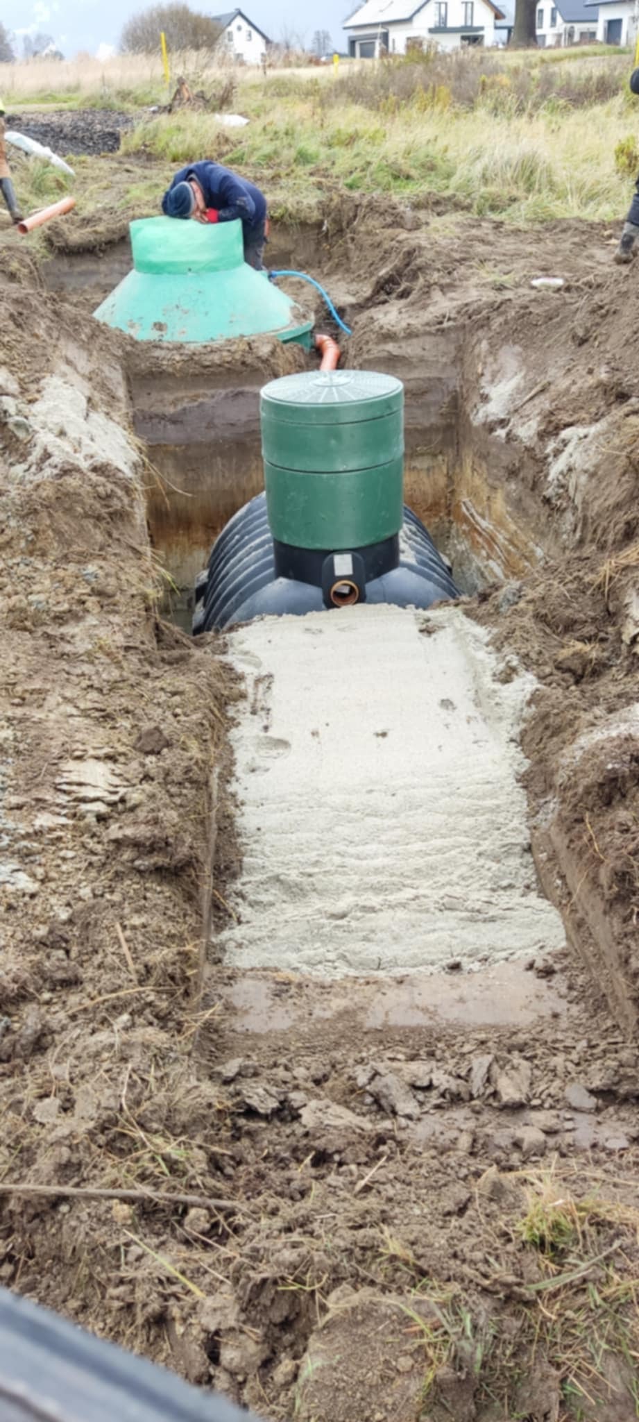 Czarna plastikowa studzienka kanalizacyjna z zieloną pokrywą umieszczona w wykopie, częściowo zasypana piaskiem, widoczny pracownik w tle.