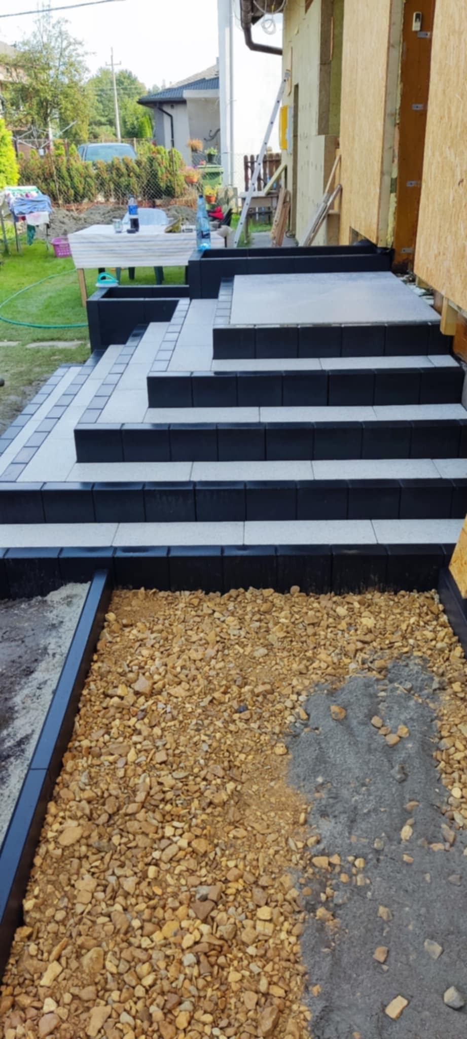 Schody wejściowe do domu w trakcie budowy, wykończone czarno-białą kostką, widoczna podsypka z kamieni i piasku. W tle trawnik i budynek.