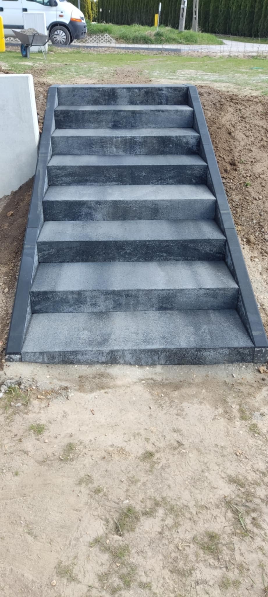Schody zewnętrzne z szarej kostki brukowej prowadzące na podwyższony teren, obrzeża wykończone ciemniejszym kolorem, w tle widoczny samochód dostawczy i wózek budowlany.