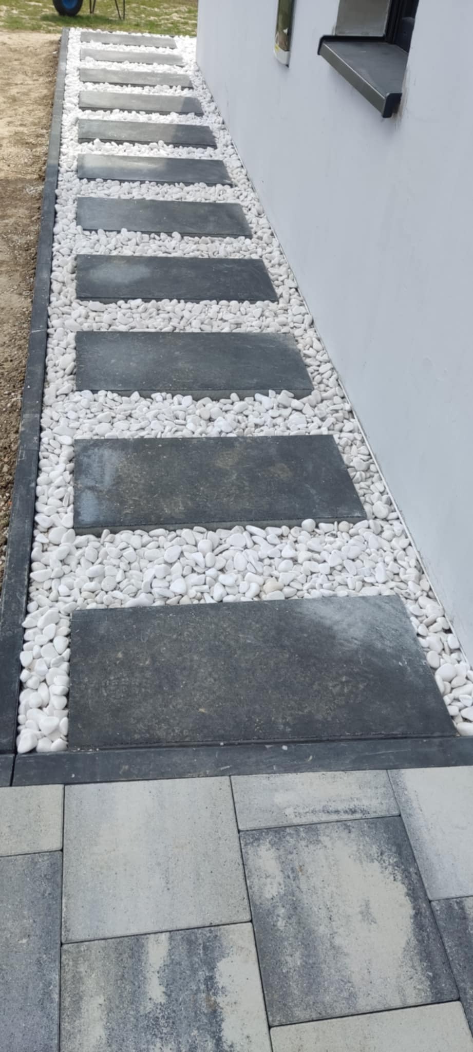 Ścieżka z ciemnych, prostokątnych płyt ułożonych w białym żwirze, obramowana ciemną kostką brukową przy białej ścianie budynku.