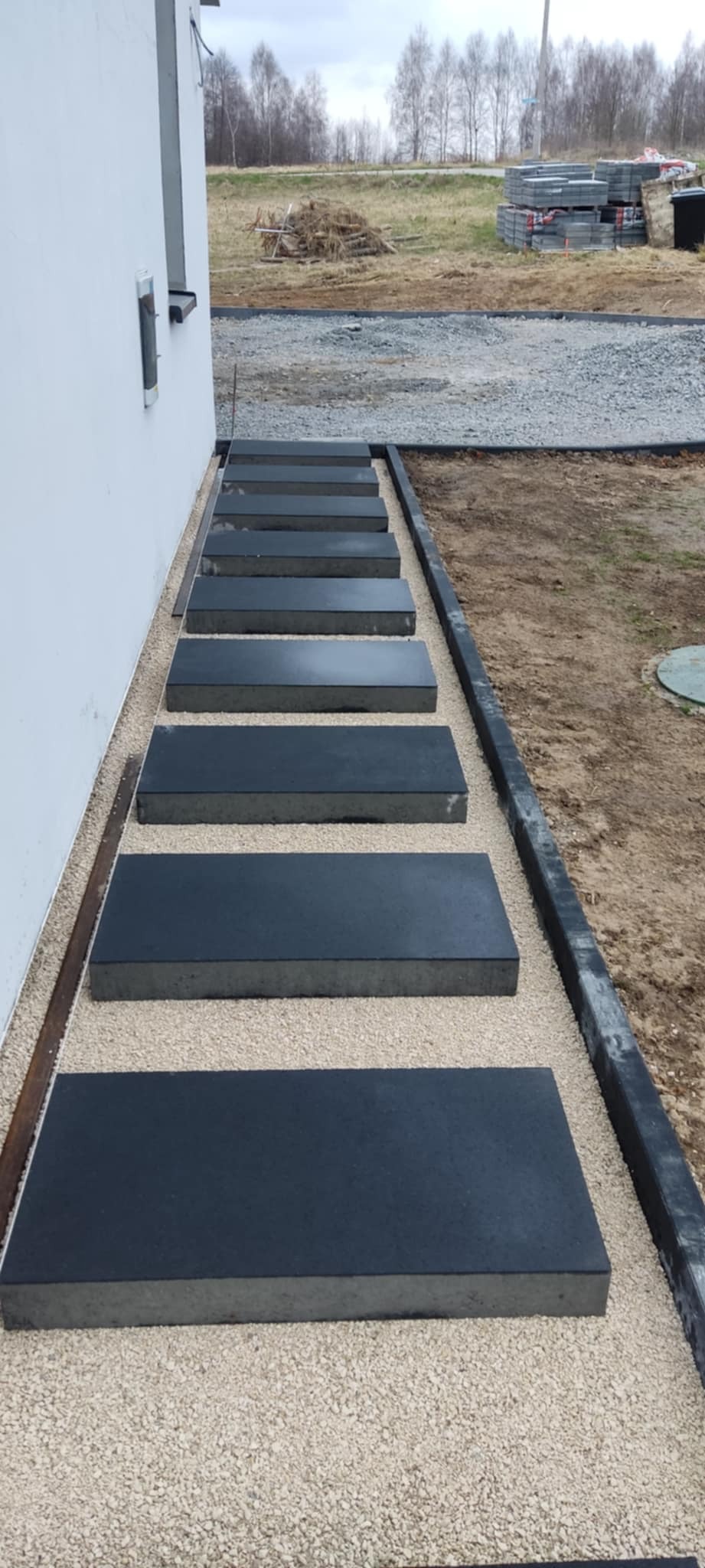 Nowoczesne, prostokątne płyty chodnikowe w kolorze grafitowym, ułożone wzdłuż białej ściany budynku na jasnym żwirze, tworzące ścieżkę. Krawężnik oddziela żwir od ziemi. W tle widoczne stosy...