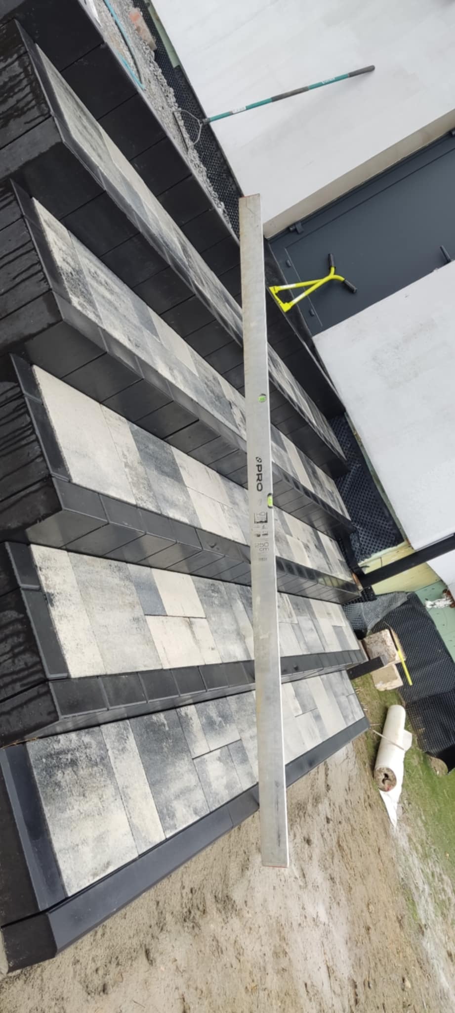 Ukończona nawierzchnia z szaro-białej kostki brukowej obramowanej czarnym obrzeżem, sprawdzana poziomicą, widoczny teren wokół i narzędzia.