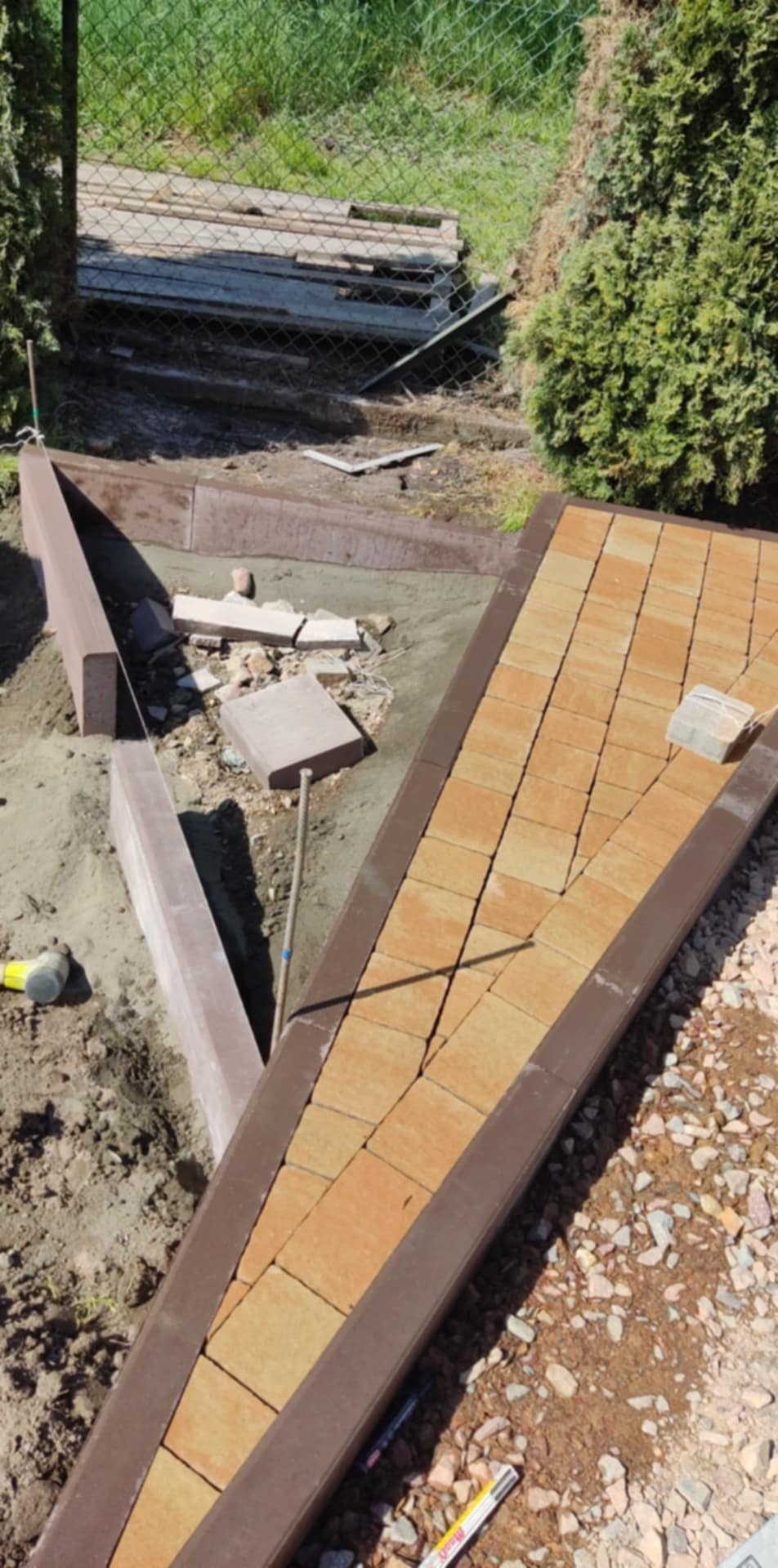 Fragment ścieżki z kostki brukowej w trakcie tworzenia, z brązowymi obrzeżami i częściowo ułożonym wzorem, narzędzia pracy widoczne obok.