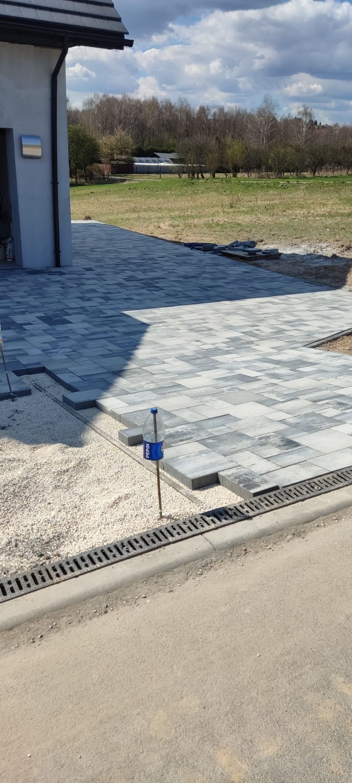 Układanie szarej kostki brukowej przed domem. Widoczny fragment ułożonej nawierzchni, podsypka z białego grysu, korytko odwadniające i butelka po napoju jako znacznik.