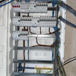 Karol Kowalewski - Rozdzielnica elektryczna w trakcie montażu: widoczne zabezpieczenia, listwy zaciskowe i przewody w różnych kolorach. Instalacja w białej obudowie, surowy mur w tle.