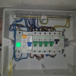 Karol Kowalewski - Rozdzielnica elektryczna w białej obudowie z widocznymi bezpiecznikami, przewodami i listwami zaciskowymi. Jeden z bezpieczników z zieloną diodą. Widoczne ślady montażu na ścianie.