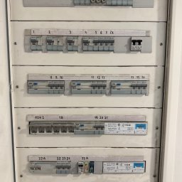 Karol Kowalewski - Rozdzielnica elektryczna z bezpiecznikami i wyłącznikami, widoczne oznaczenia numerów i parametrów. Element instalacji elektrycznej wewnątrz budynku.