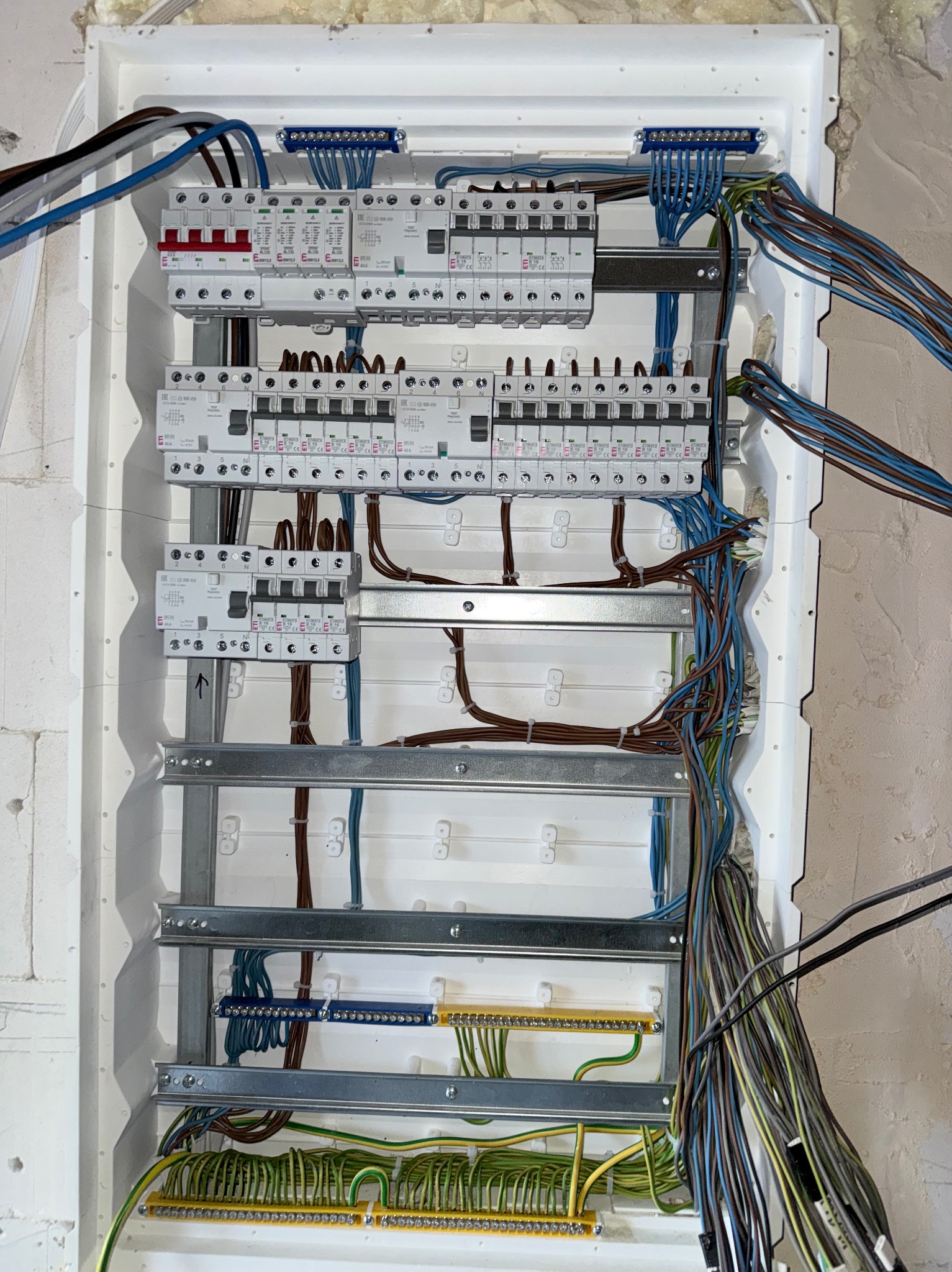 Rozdzielnica elektryczna w trakcie montażu: widoczne zabezpieczenia, listwy zaciskowe i przewody w różnych kolorach. Instalacja w białej obudowie, surowy mur w tle.