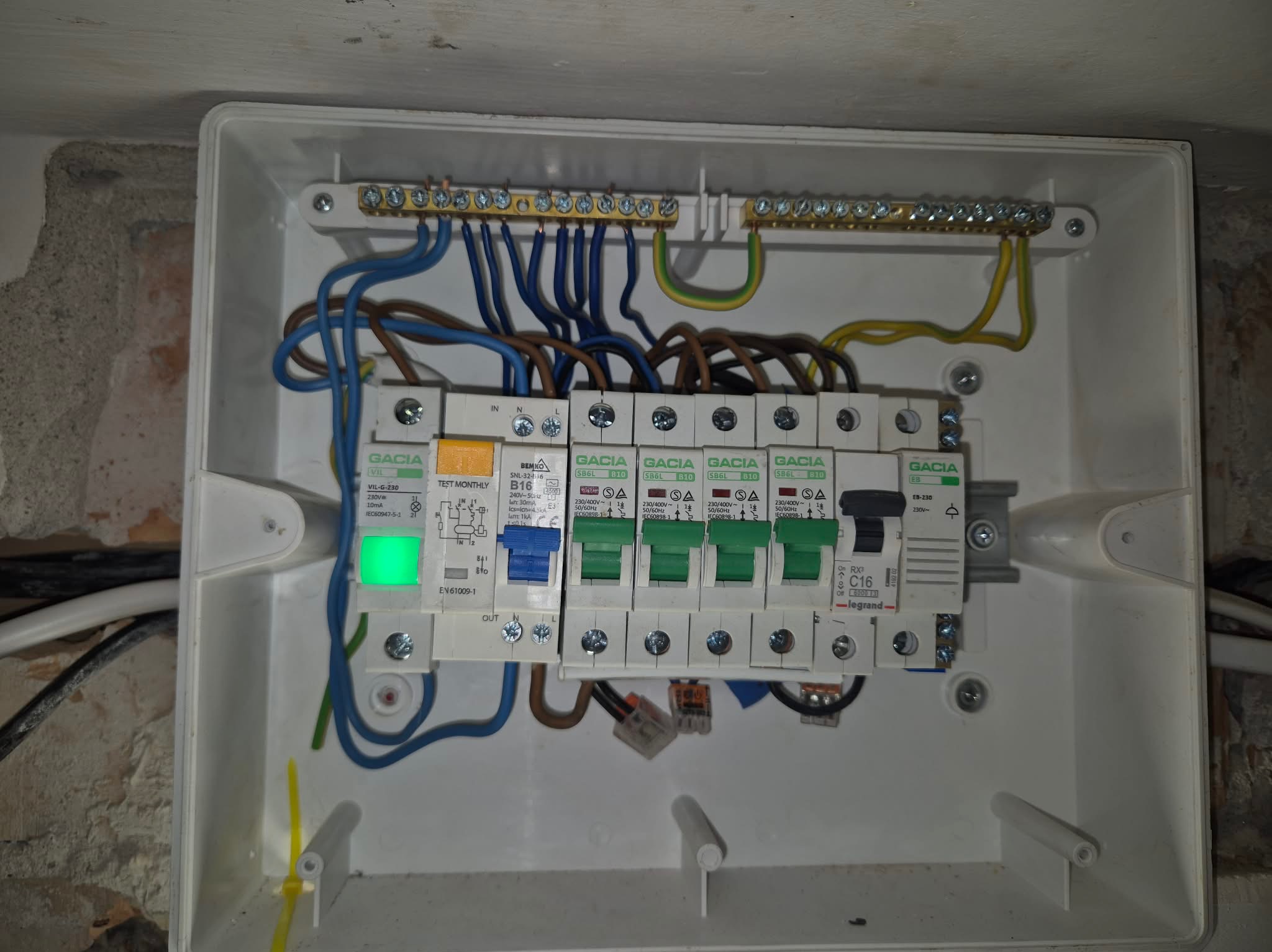 Rozdzielnica elektryczna w białej obudowie z widocznymi bezpiecznikami, przewodami i listwami zaciskowymi. Jeden z bezpieczników z zieloną diodą. Widoczne ślady montażu na ścianie.