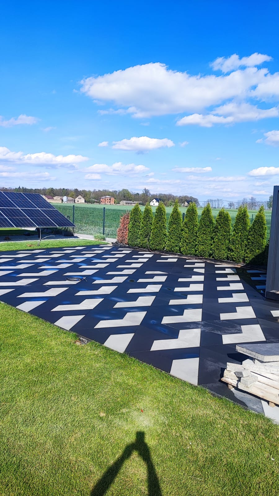 Nowoczesna, geometryczna kostka brukowa w czarno-białe wzory, otoczona zielonym trawnikiem i tujami, z panelami słonecznymi w tle, pod błękitnym niebem.