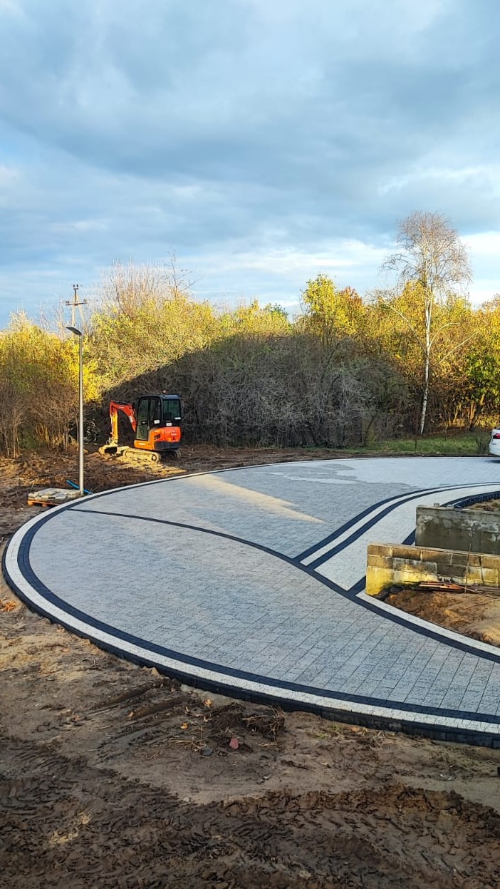 Układanie kostki brukowej w Mirkowie: Mini koparka obok nowej, szarej kostki brukowej z czarną obwódką, tworzącej elegancki podjazd. Prace ziemne w toku.