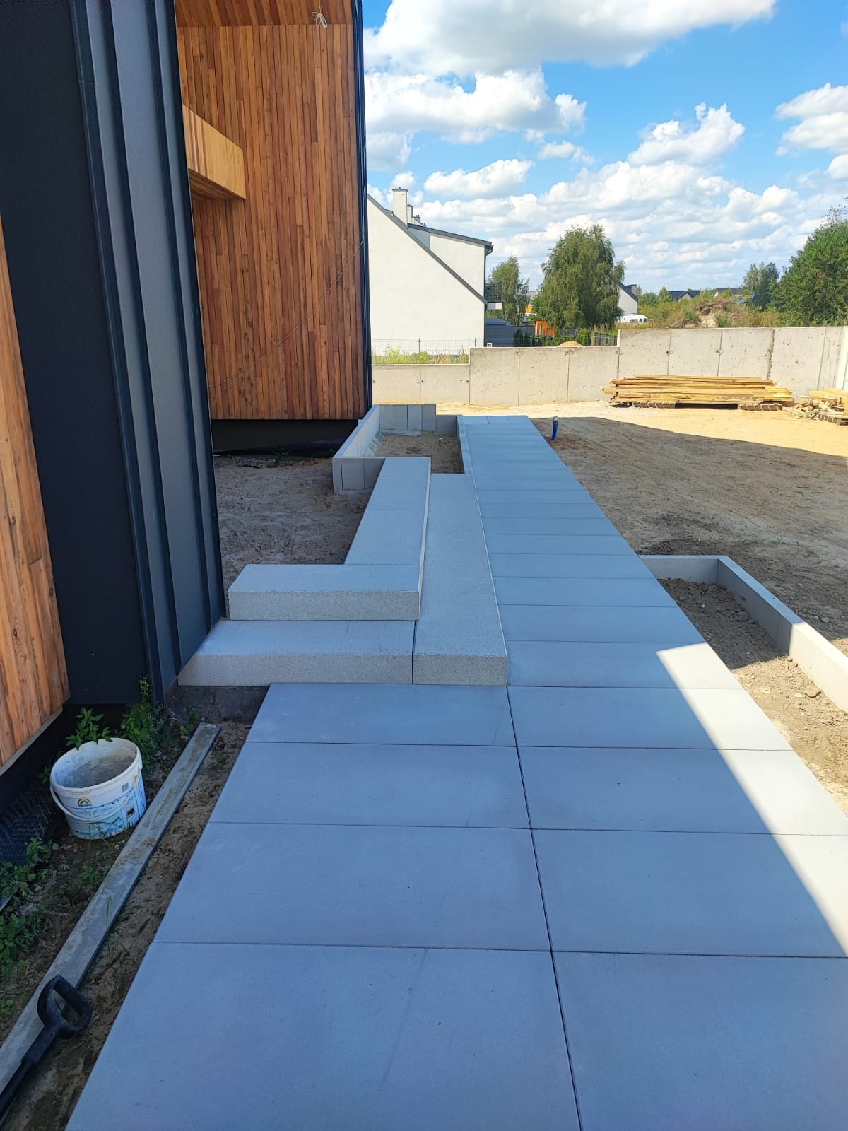 Nowoczesna ścieżka z szarych płyt prowadząca do domu z drewnianą elewacją. Widoczne schody wejściowe i przygotowane rabaty. Słoneczny dzień.