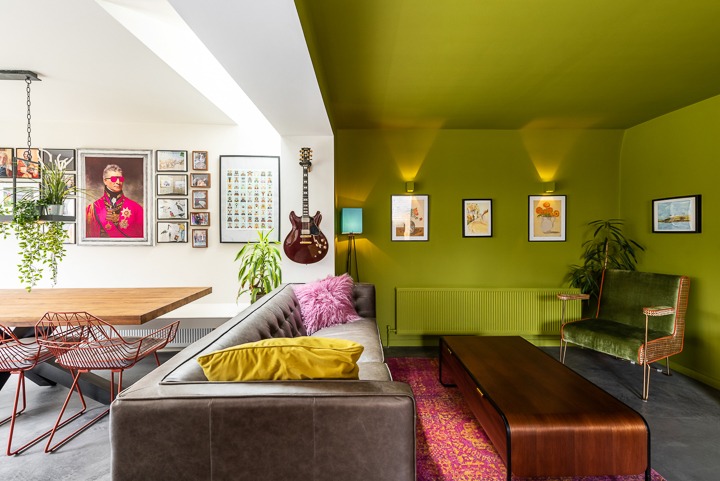 Salon z zieloną ścianą i sufitem, brązową sofą z kolorowymi poduszkami, drewnianym stołem i gitarą na ścianie. Nietypowy wystrój wnętrza.