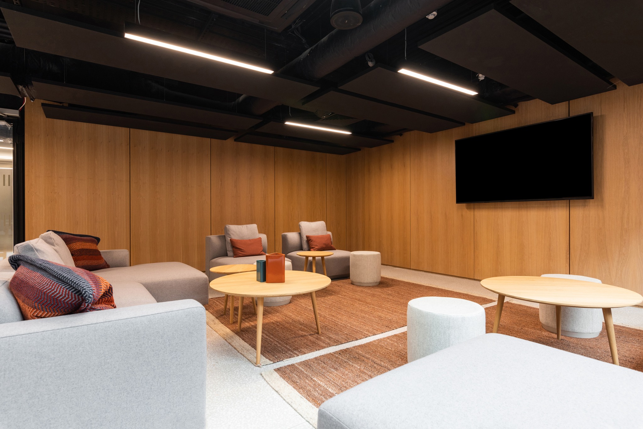 Nowoczesne wnętrze biurowe z drewnianymi panelami, szarymi sofami i okrągłymi stolikami. Minimalistyczny design, oświetlenie LED i duży telewizor na ścianie.