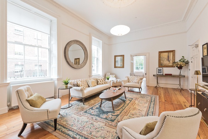 Jasne wnętrze salonu z drewnianą podłogą, dywanem w kwiaty i beżowymi meblami w stylu klasycznym. Na ścianach wiszą obrazy i lustro.