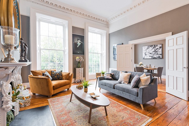 Elegancki salon z szarą sofą, musztardowym fotelem i drewnianą podłogą. Dekoracyjny kominek, duże okna i stylowe dodatki tworzą przytulną atmosferę.