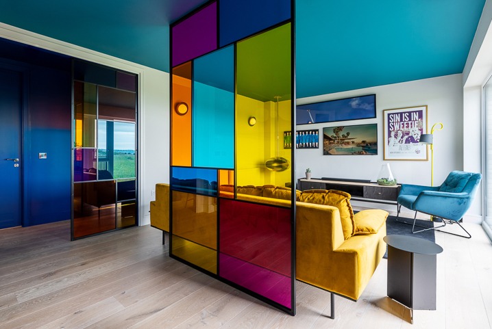 Kolorowa szklana ścianka działowa w nowoczesnym salonie z żółtą sofą i niebieskimi akcentami. Widok na drzwi i okno z zielonym krajobrazem.