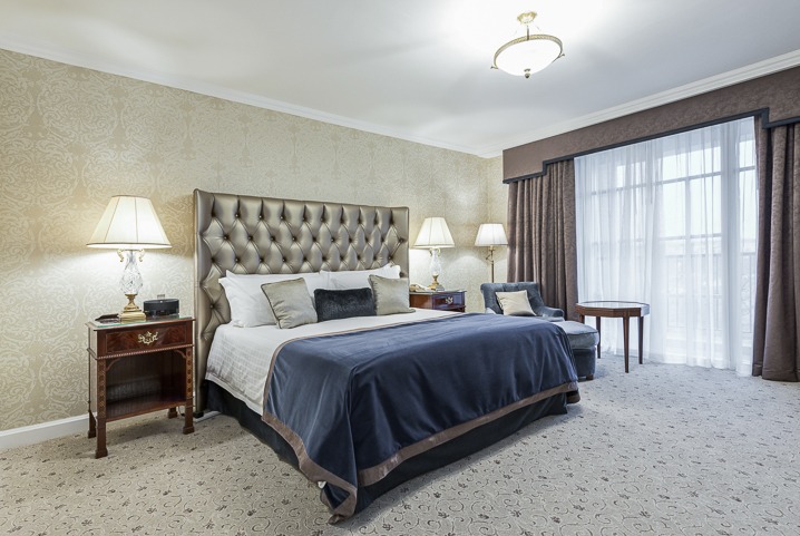 Elegancka sypialnia z pikowanym zagłówkiem, ciemnoniebieską narzutą i wzorzystą tapetą. Na stoliku nocnym kryształowa lampka i zegar.