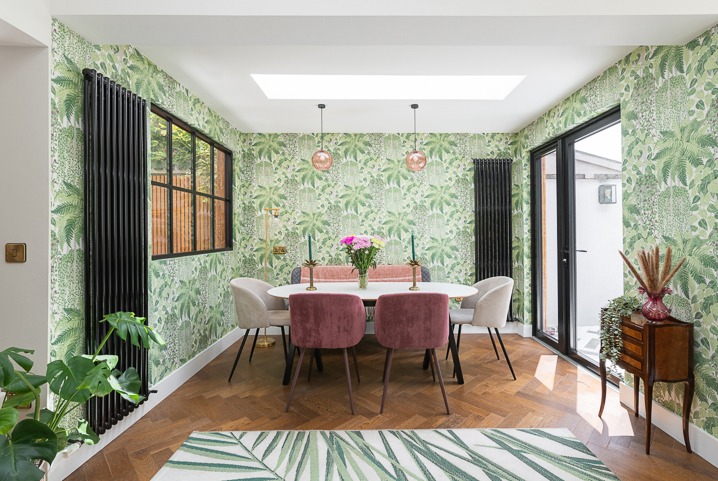 Jadalnia z owalnym stołem, welurowymi krzesłami i ścianami w tropikalne wzory. Na stole kwiaty, a nad nim lampy w stylu vintage. Parkiet w jodełkę i dywan z motywem liści.