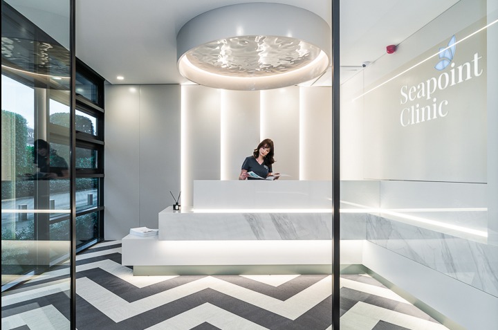 Nowoczesne wnętrze kliniki z recepcją, oświetleniem LED i wzorzystą podłogą. Kobieta w recepcji przegląda dokumenty. Widoczne logo 'Seapoint Clinic'.