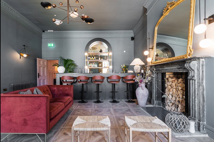 Stylowe wnętrze z barkiem i czerwonym welurowym narożnikiem. Nad barkiem półka z oświetleniem i alkoholem. Kominek z lustrem w złotej ramie i dekoracyjnym drewnem.
